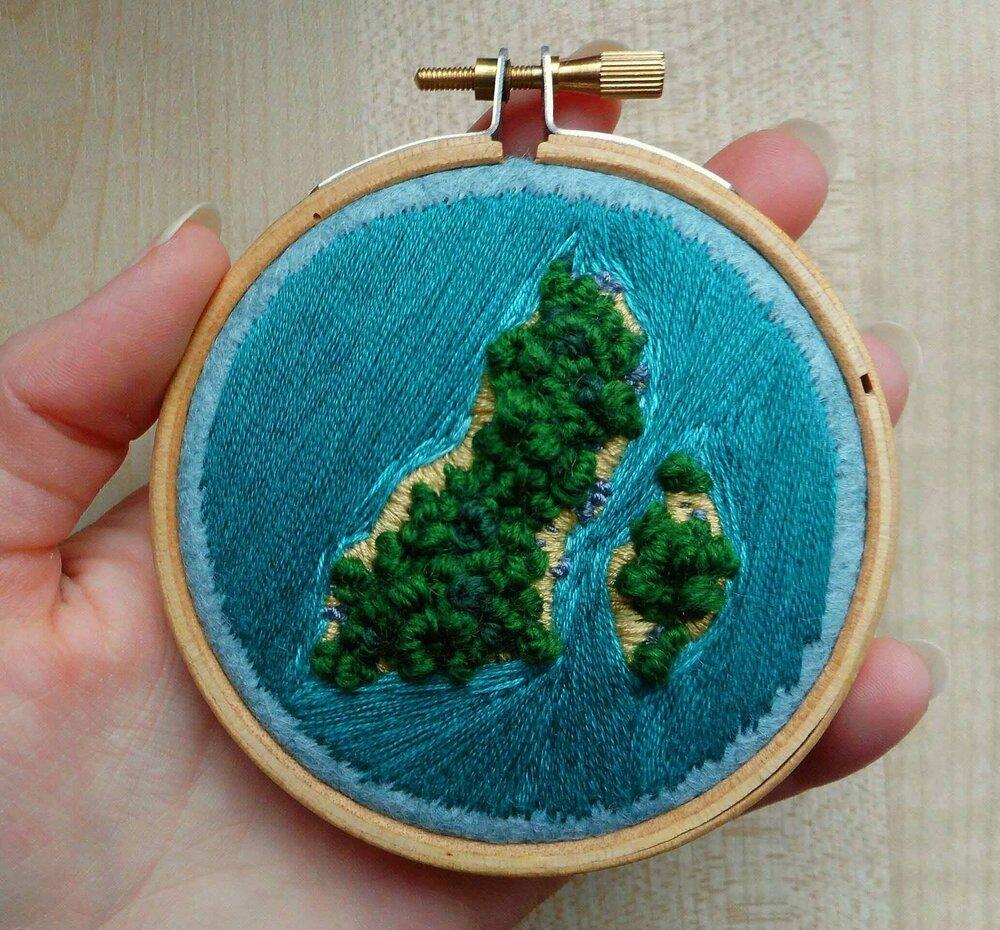 Small embroidery hoop depicting an aerial view of a lush green island at sea