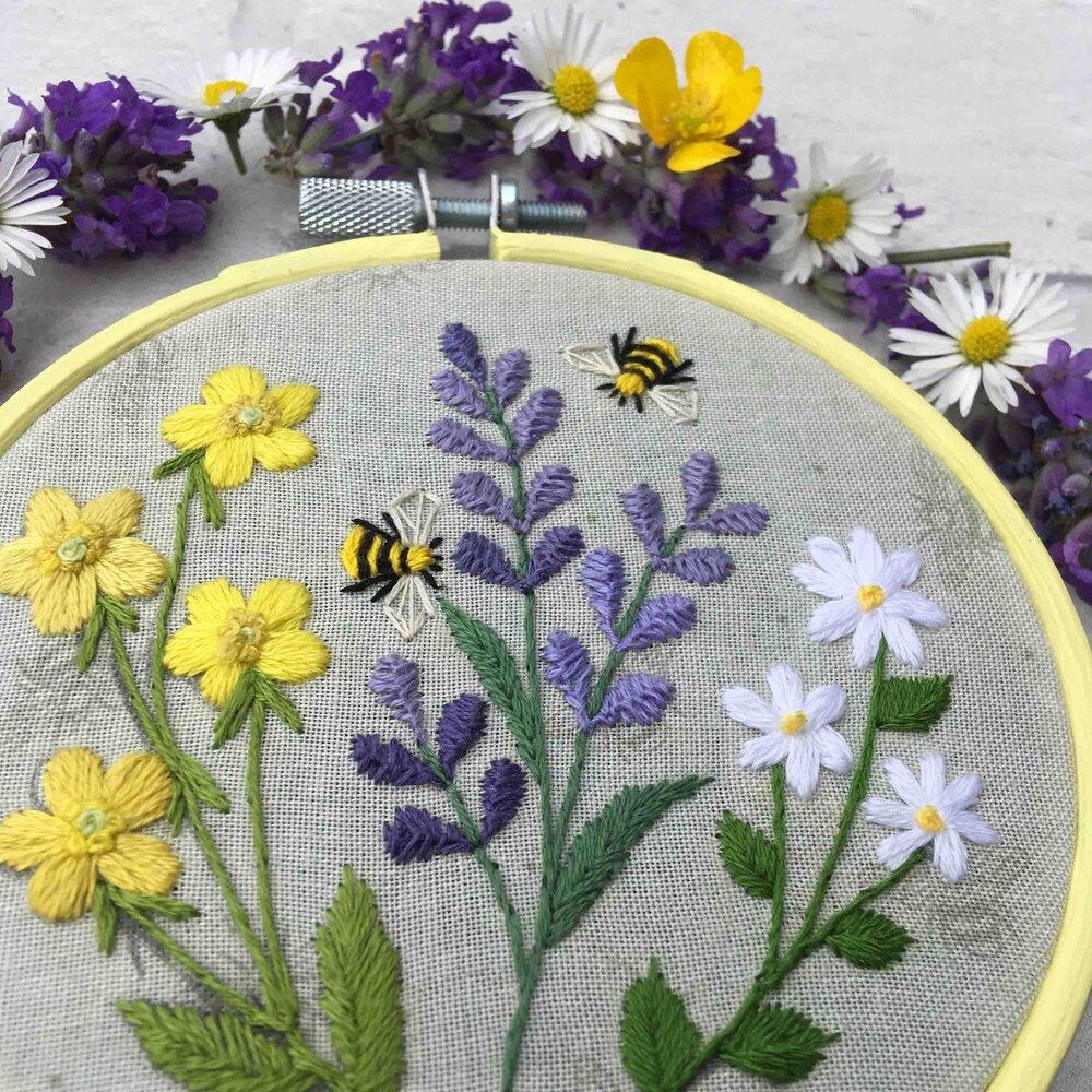 Yellow, purple, and white flowers embroidered onto grey fabric. Two bees surround the flowers.