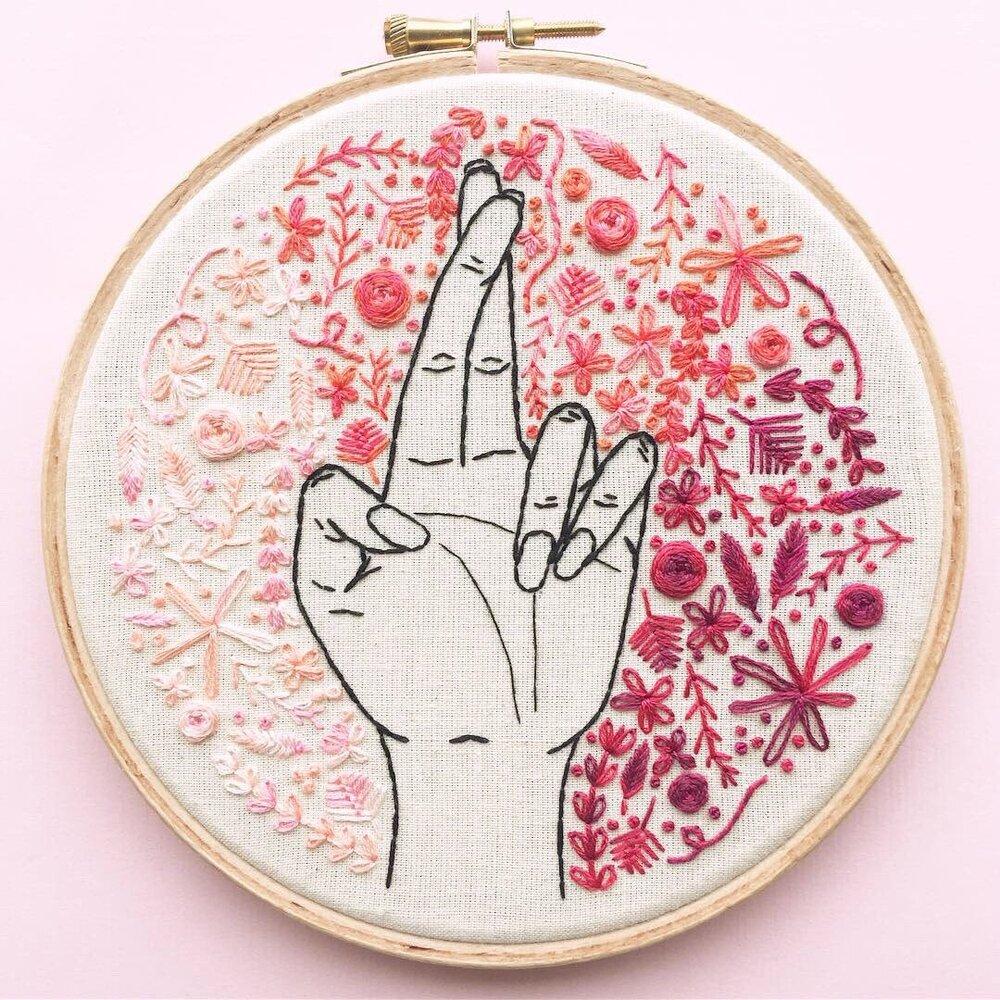 Embroidery hoop with a crossed finger design, surrounded by embroidered flowers in autumnal colours