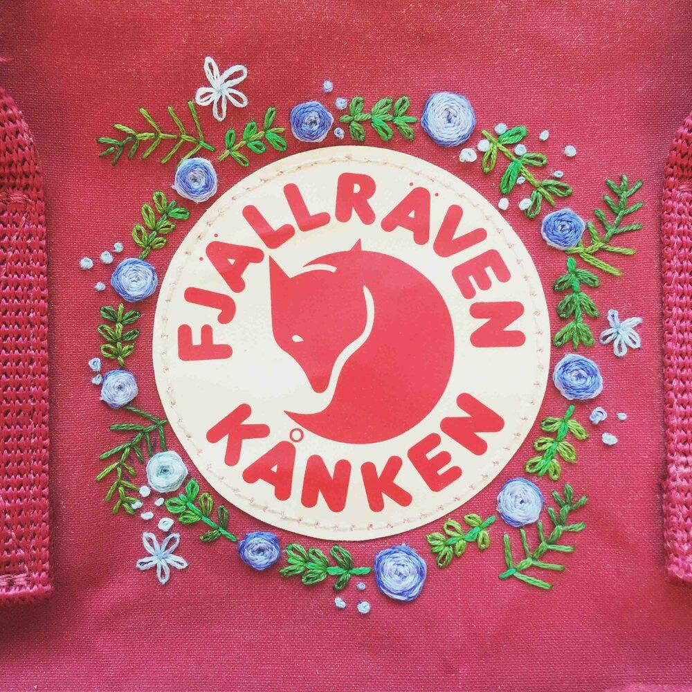 Image depicting the Fjallraven Kanken bag logo with small, embroidered flowers and greenery around it.